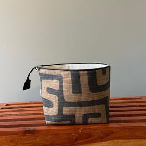 Pode incluir: Uma bolsa retangular com fecho de correr em tecido texturizado com um padr&atilde;o geom&eacute;trico repetitivo em cinzento-carv&atilde;o escuro sobre um fundo bege. A bolsa tem um fecho preto com um puxador de tecido preto e um forro interior branco.