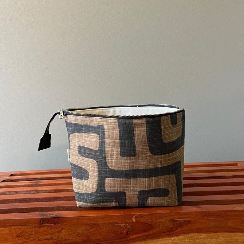 Pode incluir: Uma bolsa retangular com fecho de correr em tecido texturizado com um padr&atilde;o geom&eacute;trico repetitivo em cinzento-carv&atilde;o escuro sobre um fundo bege. A bolsa tem um fecho preto com um puxador de tecido preto e um forro interior branco.