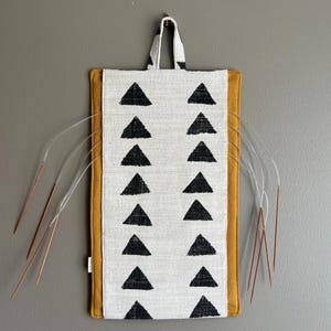 May include: A rectangular knitting needle case with a white background and black triangle pattern. The case has a mustard yellow border and a matching handle. Several knitting needles are visible.