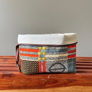 May include: A rectangular fabric pouch featuring a geometric pattern of red, yellow, blue, and black grid lines. The top section is a solid off-white canvas fabric with a brown zipper closure along the top edge and a brown side pull tab.