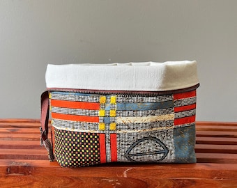 May include: A rectangular fabric pouch featuring a geometric pattern of red, yellow, blue, and black grid lines. The top section is a solid off-white canvas fabric with a brown zipper closure along the top edge and a brown side pull tab.