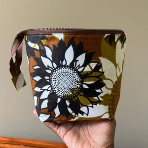 May include: A zippered pouch featuring a bold floral pattern with large black, white, and mustard yellow sunflower-like petals against a brown background. The bag has a brown zipper closure along the top edge and a small brown fabric pull tab.