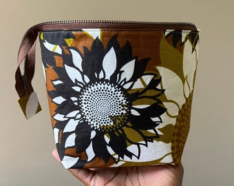 Pode incluir: Uma bolsa com fecho de correr apresentando um padrão floral arrojado com grandes pétalas semelhantes a girassóis em preto, branco e amarelo mostarda sobre um fundo castanho. O saco tem um fecho castanho no topo e uma pequena aba de tecido.