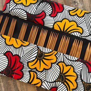 Könnte beinhalten: Eine schwarze Stricknadel-Tasche mit einem farbenfrohen Blumenmuster in Rot, Gelb und Weiß. Die Tasche ist geöffnet und zeigt mehrere Stricknadeln aus Holz. Die Nadeln sind ordentlich angeordnet und einsatzbereit.