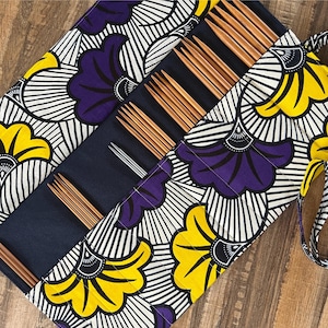 May include: A fabric knitting needle case with a vibrant floral pattern in yellow, purple, and white. The case is open, revealing several wooden knitting needles and a silver crochet hook. The case has a navy blue interior.
