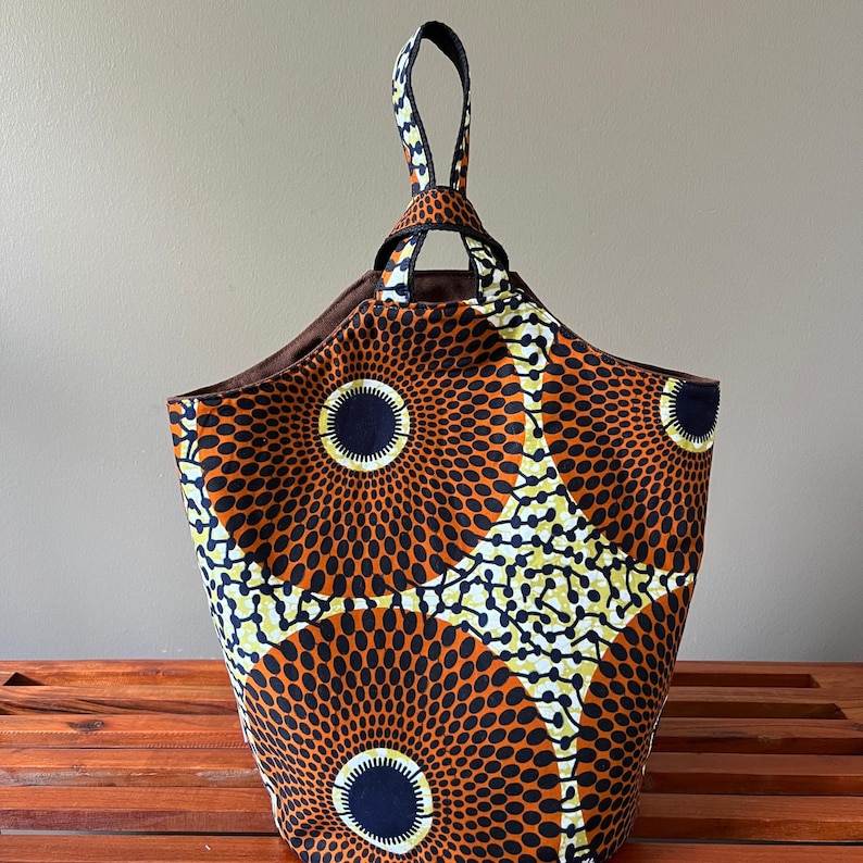 May include: A fabric tote bag with a vibrant African print. The bag features a repeating pattern of orange, black, and yellow circles and dots. It has a brown interior and a handle made of the same patterned fabric.