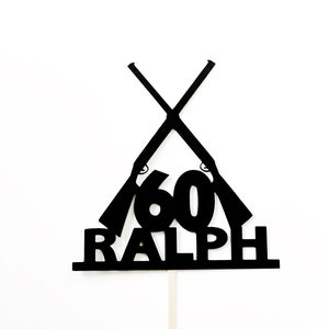 May include: Black silhouette cake topper with two crossed rifles, the number 60, and the name Ralph.