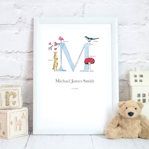 May include: A white framed print featuring a blue letter M surrounded by illustrations of a magpie, a mushroom, a flower, and a meerkat. The print includes the name "Michael James Smith" and the date "01.01.2020".