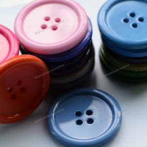 以下が含まれることがあります： 4つの穴のあるカラフルなプラスチックボタンの詰め合わせ。ボタンはピンク、オレンジ、ブルー、グリーンなど、さまざまな色があります。