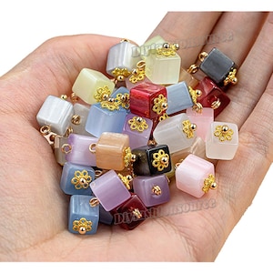 Op de afbeelding: Een verzameling kleurrijke, vierkante bedels met gouden accenten. De bedels zijn gemaakt van verschillende materialen, waaronder glas, keramiek en metaal. Ze zijn perfect om een ​​toets van whimsy toe te voegen aan sieraden, ambachten en andere projecten.