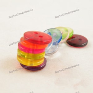 Puede incluir: Una colección de botones translúcidos de colores en varios tonos, incluyendo rojo, naranja, rosa, amarillo y morado. Los botones están apilados y dispuestos en fila, mostrando su diseño de dos agujeros. Los botones son de plástico.