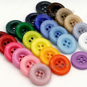 Peut inclure: Assortiment de 16 boutons en plastique colorés avec quatre trous chacun. Les boutons sont disposés en cercle, les couleurs allant du rouge au violet.