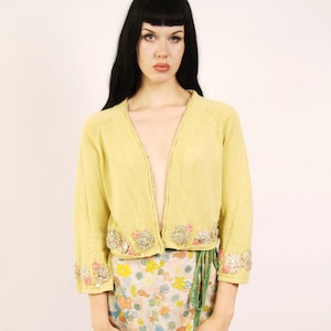 May include: A light yellow cardigan with floral embellishments on the cuffs and hem. The outfit includes a floral print wrap skirt with a green trim. Brown open-toe sandals complete the look.