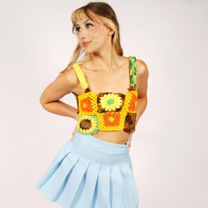 May include: A cropped crochet top featuring a vibrant sunflower and square pattern in shades of yellow, orange, and green with thick straps, paired with a light blue pleated mini skirt and brown leather open-toe clogs with wooden soles.