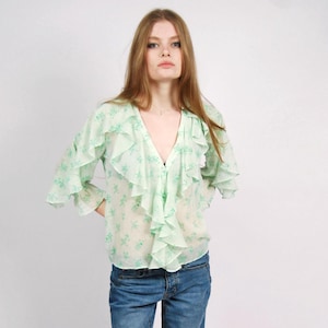 May include: A pale green floral blouse with frill detailing, worn with blue denim jeans. The blouse features a V-neck and long sleeves. The jeans are a straight-leg cut. White trainers complete the look.