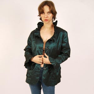 May include: A dark green jacket with a ruffled collar and button closure. The jacket is paired with blue jeans. The outfit is completed with brown sandals.