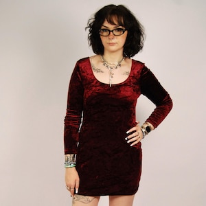 May include: A long-sleeved, deep burgundy velvet mini dress featuring a scoop neckline and a fitted silhouette. The fabric has a crushed texture that catches the light, creating subtle variations in the dark red colour across the garment.