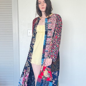 Puede incluir: Un abrigo de patchwork de manga larga y largo hasta la rodilla con una variedad de patrones de tela que incluyen estampados florales, diseños geométricos y colores sólidos en tonos rojo, azul, negro y amarillo, sobre un vestido corto amarillo.