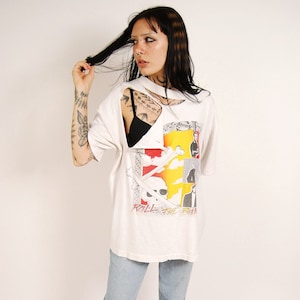 May include: A white t-shirt with a graphic print of a skull and crossbones and the text "Roll the Bones". The shirt has a large hole in the front and is being worn with light blue jeans and pink slides.