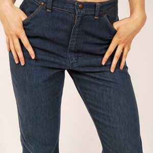 May include: Dark blue denim jeans with a classic five-pocket design. The jeans have a button and zipper closure, and the model is wearing a light purple top. The jeans are a straight-leg style.
