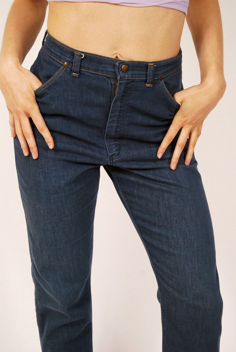 May include: Dark blue denim jeans with a classic five-pocket design. The jeans have a button and zipper closure, and the model is wearing a light purple top. The jeans are a straight-leg style.