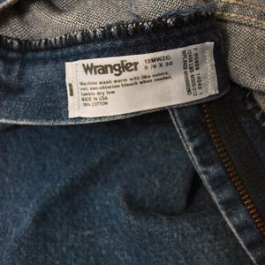 Op de afbeelding: Close-up van een blauw denim kledingstuk met een wit label. Het label leest "Wrangler" met maat informatie en was instructies. Het kledingstuk is gemaakt van 100% katoen.