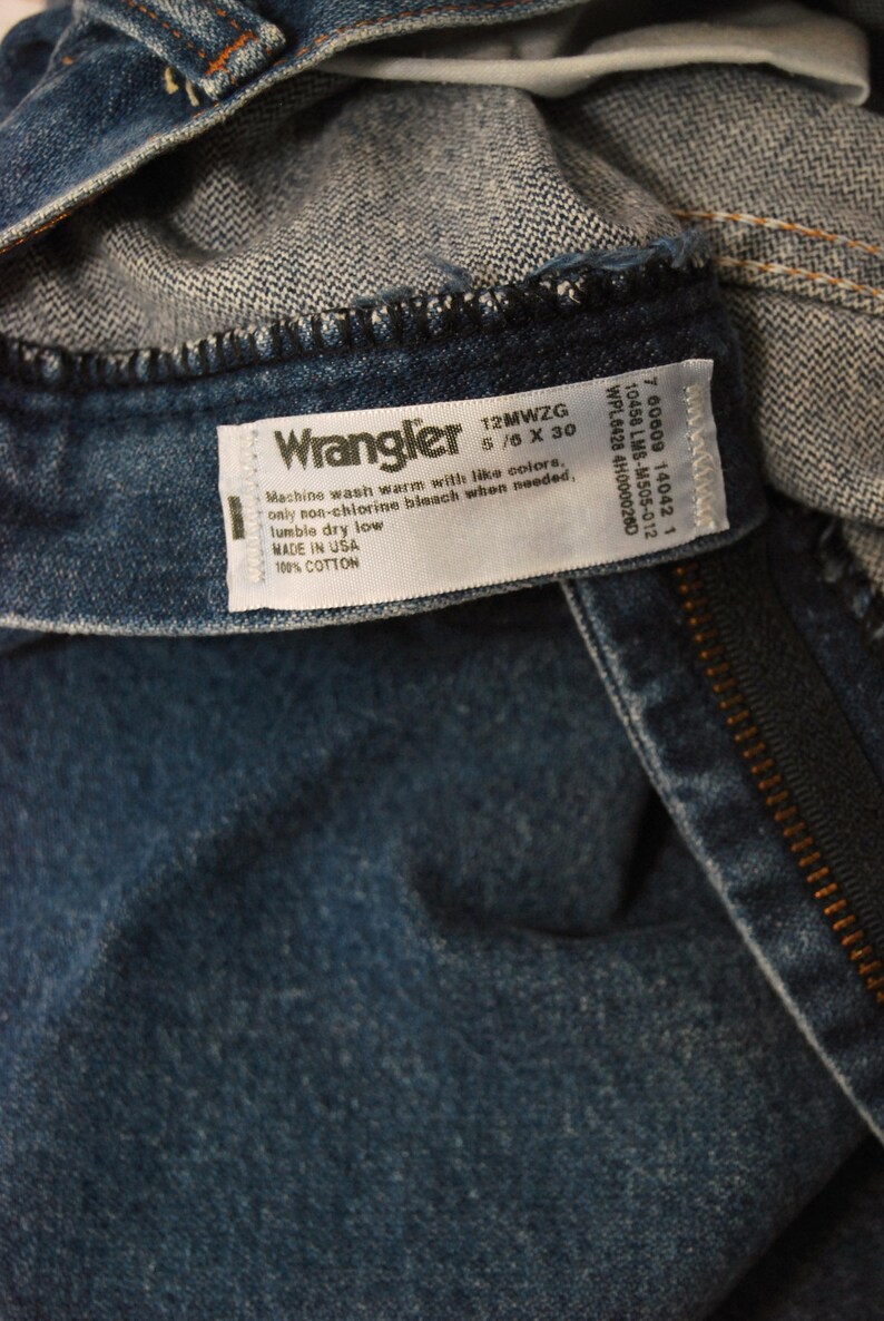 Op de afbeelding: Close-up van een blauw denim kledingstuk met een wit label. Het label leest "Wrangler" met maat informatie en was instructies. Het kledingstuk is gemaakt van 100% katoen.