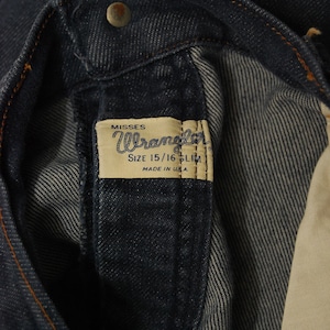 May include: Close-up of dark blue denim jeans. The jeans feature a metal button and a label that reads "Misses Wrangler SIZE 15/16 SLIM MADE IN U.S.A."