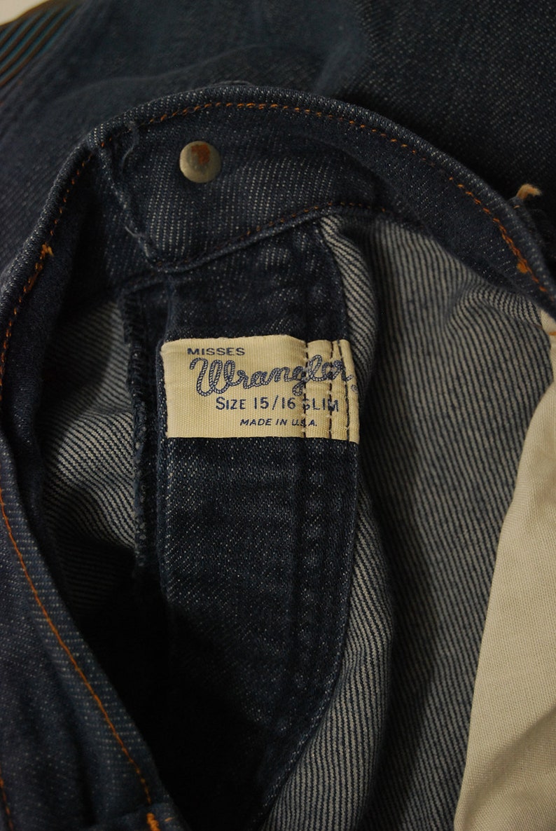 May include: Close-up of dark blue denim jeans. The jeans feature a metal button and a label that reads "Misses Wrangler SIZE 15/16 SLIM MADE IN U.S.A."