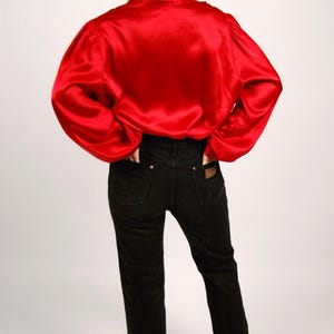 May include: A red satin blouse with long sleeves and black denim jeans. The blouse has a high collar and the jeans have a classic five-pocket design. The model is wearing black heels.