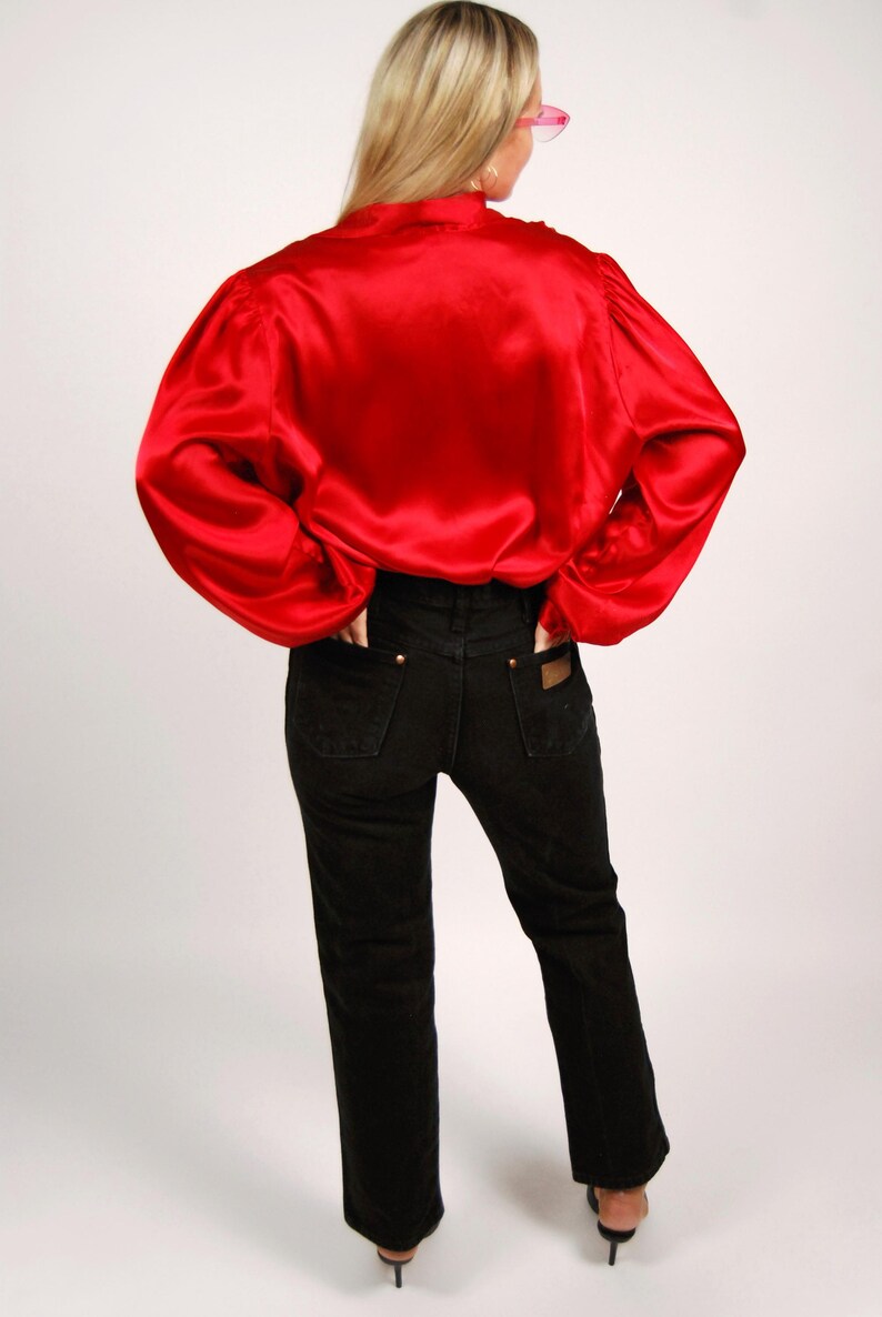 May include: A red satin blouse with long sleeves and black denim jeans. The blouse has a high collar and the jeans have a classic five-pocket design. The model is wearing black heels.