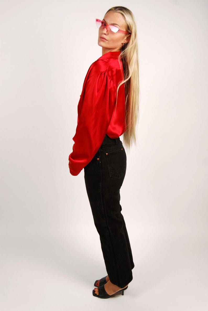 May include: A red satin blouse with long sleeves and black denim jeans. The outfit is paired with black heeled sandals and pink-framed sunglasses. The model is facing away from the camera.