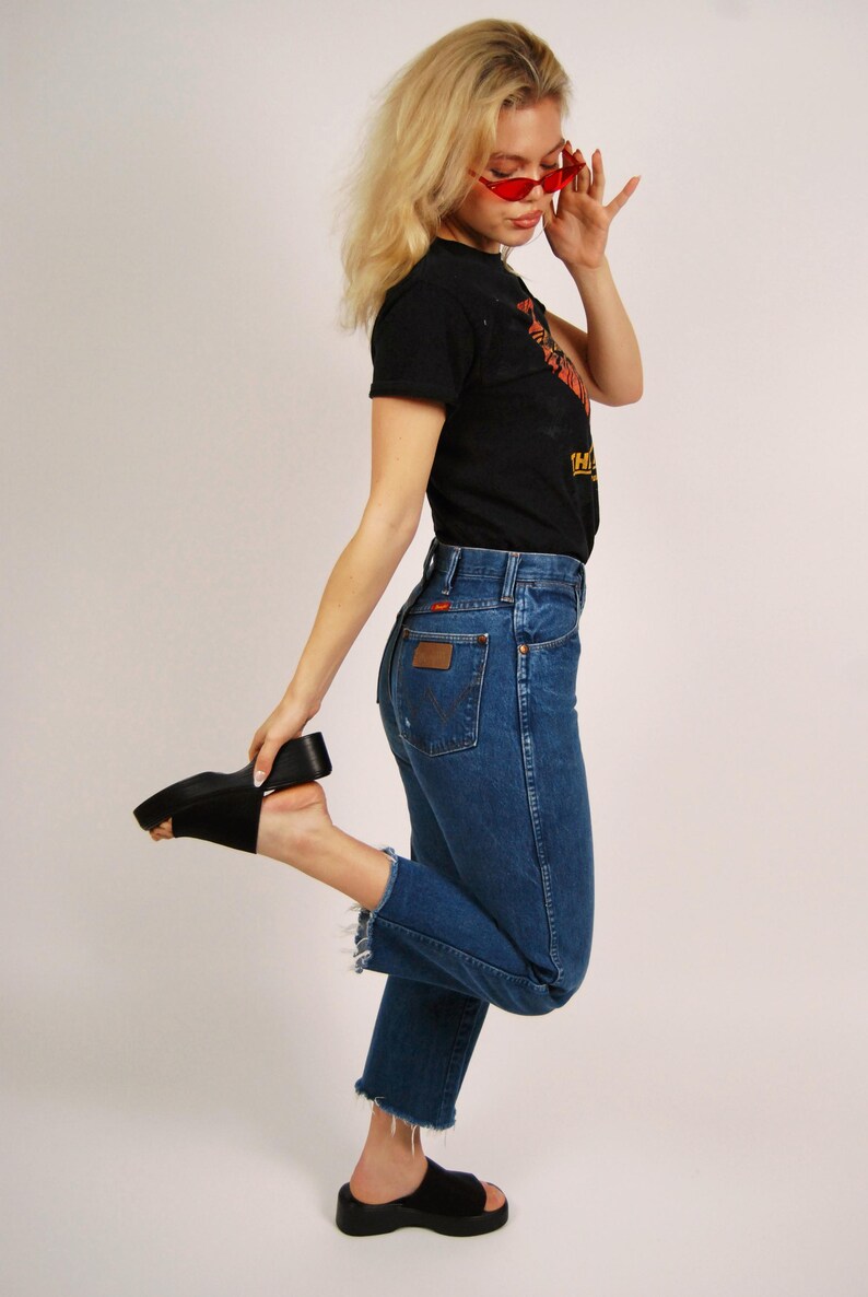 Op de afbeelding: Een persoon draagt een zwart t-shirt, een blauwe spijkerbroek en zwarte plateausandalen. De jeans heeft een hoge taille en een gerafelde zoom. De persoon draagt ook een rode zonnebril.