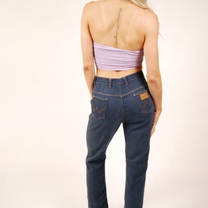 May include: A woman wearing a light purple tube top and dark blue denim jeans. The jeans have a brown leather patch on the back pocket. Black platform sandals complete the outfit. The woman has blonde hair.