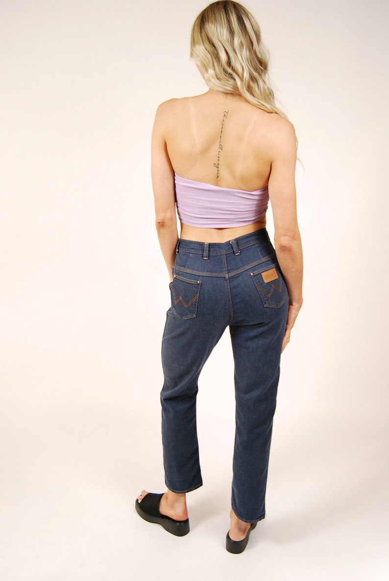 May include: A woman wearing a light purple tube top and dark blue denim jeans. The jeans have a brown leather patch on the back pocket. Black platform sandals complete the outfit. The woman has blonde hair.