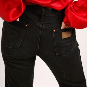 May include: Black denim jeans with a classic five-pocket design. The jeans have a straight leg cut and feature copper-colored rivets and a leather patch on the back pocket. A red satin blouse is visible.