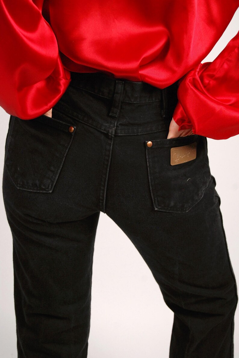 May include: Black denim jeans with a classic five-pocket design. The jeans have a straight leg cut and feature copper-colored rivets and a leather patch on the back pocket. A red satin blouse is visible.