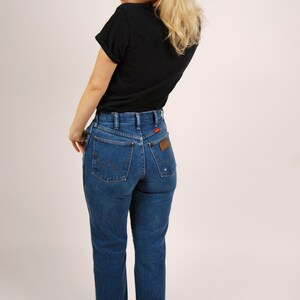 Op de afbeelding: Een persoon draagt een zwart t-shirt en een blauwe spijkerbroek. De jeans heeft een hoge taille en een rechte pijp. De jeans heeft een bruin leren label op de achterzak. De persoon draagt ook zwarte schoenen en een rode zonnebril.