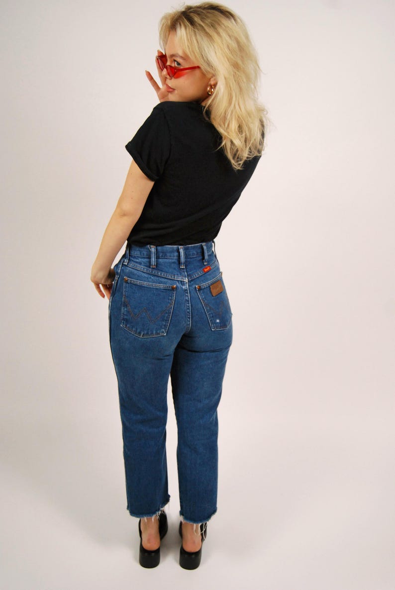 Op de afbeelding: Een persoon draagt een zwart t-shirt en een blauwe spijkerbroek. De jeans heeft een hoge taille en een rechte pijp. De jeans heeft een bruin leren label op de achterzak. De persoon draagt ook zwarte schoenen en een rode zonnebril.