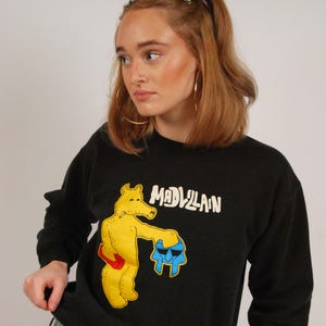 以下が含まれることがあります： 黄色の漫画のクマが青と黒の物体を抱えている黒いスウェットシャツ。スウェットシャツには白い文字で「MADVILLAIN」と印刷されています。