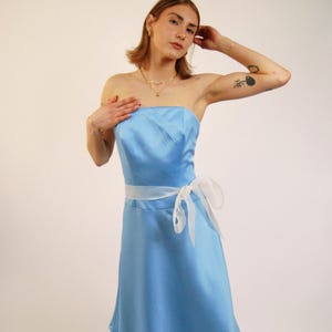 Könnte beinhalten: Ein trägerloses, hellblaues Kleid mit einem weißen Band, das um die Taille gebunden ist. Das Kleid hat einen fließenden Rock und ein eng anliegendes Oberteil. Das weiße Band ist zu einer Schleife an der Seite gebunden. Das Kleid ist aus glänzendem, satinartigem Material.