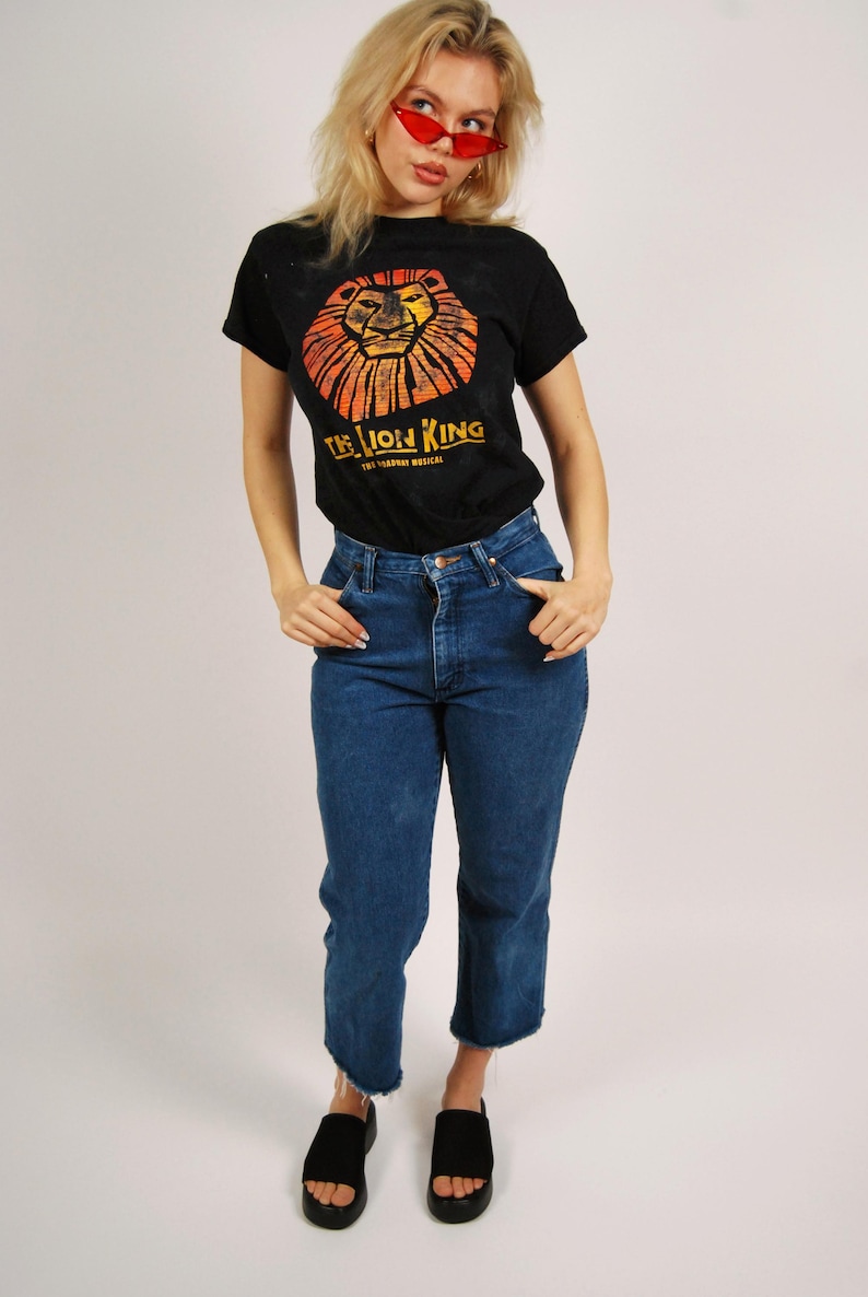 Op de afbeelding: Zwart T-shirt met een oranje en gele leeuwenafbeelding en de tekst "THE LION KING". De outfit omvat een blauwe jeans en zwarte sandalen. De persoon draagt een rode zonnebril. Witte achtergrond.