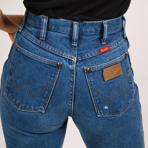 Op de afbeelding: Blauwe denim jeans met een hoge taille en een klassiek Wrangler-ontwerp. Kenmerken zijn koperen klinknagels, een leren patch en het Wrangler-logo. De jeans hebben een medium wassing en een vintage esthetiek.