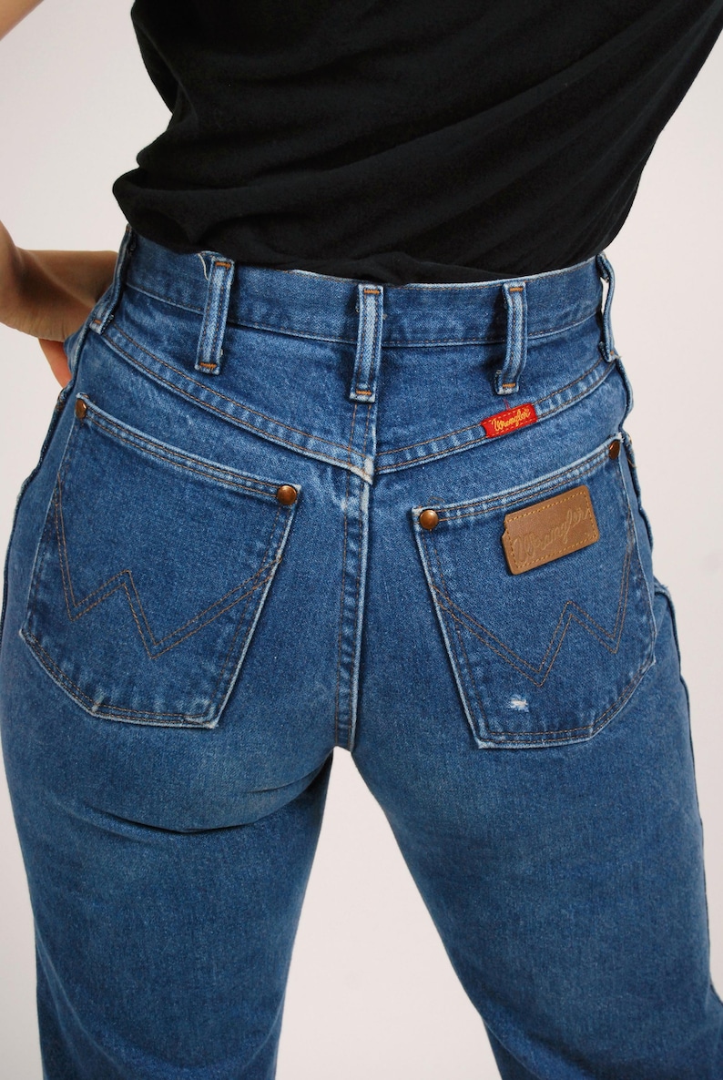 Op de afbeelding: Blauwe denim jeans met een hoge taille en een klassiek Wrangler-ontwerp. Kenmerken zijn koperen klinknagels, een leren patch en het Wrangler-logo. De jeans hebben een medium wassing en een vintage esthetiek.