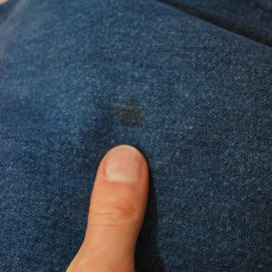 Op de afbeelding: Close-up van blauwe denimstof met een donkere vlek. De denim heeft een textuur. Een duim staat op de voorgrond.
