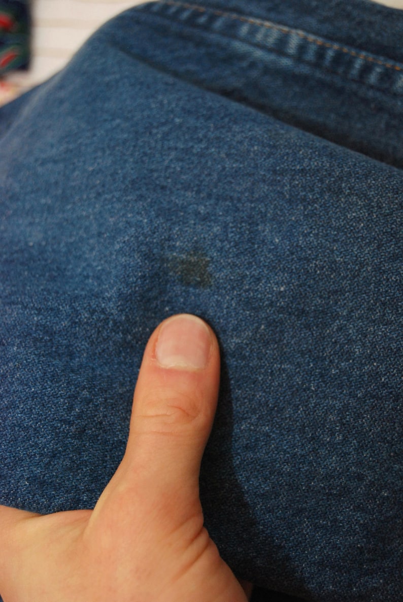 Op de afbeelding: Close-up van blauwe denimstof met een donkere vlek. De denim heeft een textuur. Een duim staat op de voorgrond.