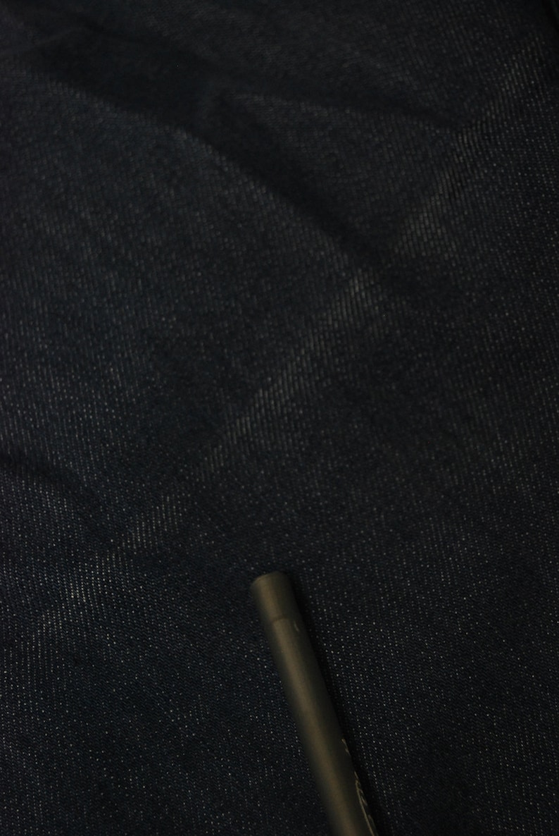 May include: Close-up of dark blue denim fabric with a subtle texture. A dark gray pen is in the lower right corner, providing a sense of scale and contrast against the denim.