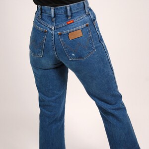 Op de afbeelding: Blauwe denim jeans met een hoge taille en een rechte pijp. De jeans hebben een Wrangler-patch op de achterzak en een rood Wrangler-label. De zoom is gerafeld.