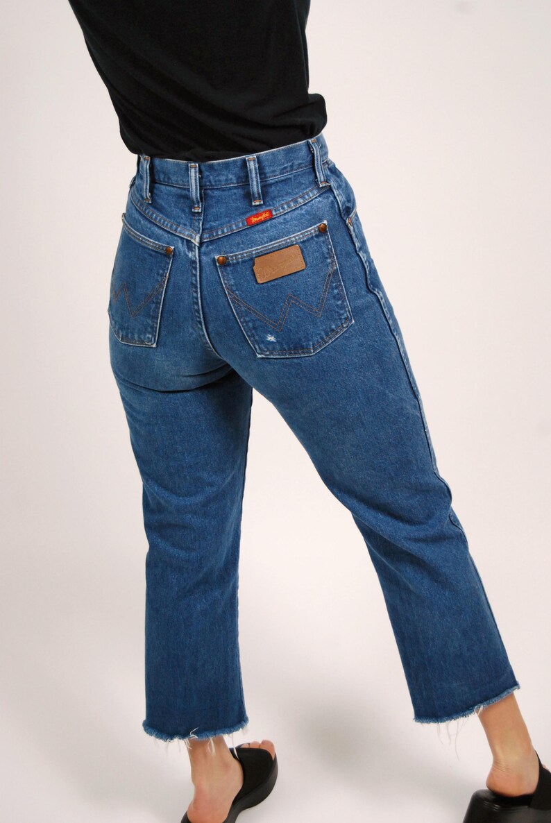 Op de afbeelding: Blauwe denim jeans met een hoge taille en een rechte pijp. De jeans hebben een Wrangler-patch op de achterzak en een rood Wrangler-label. De zoom is gerafeld.