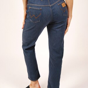 May include: Dark blue denim jeans with a high waist and straight leg. Features include orange stitching, a brown leather patch, and the Wrangler logo on the back pocket. The jeans are worn with black platform sandals.